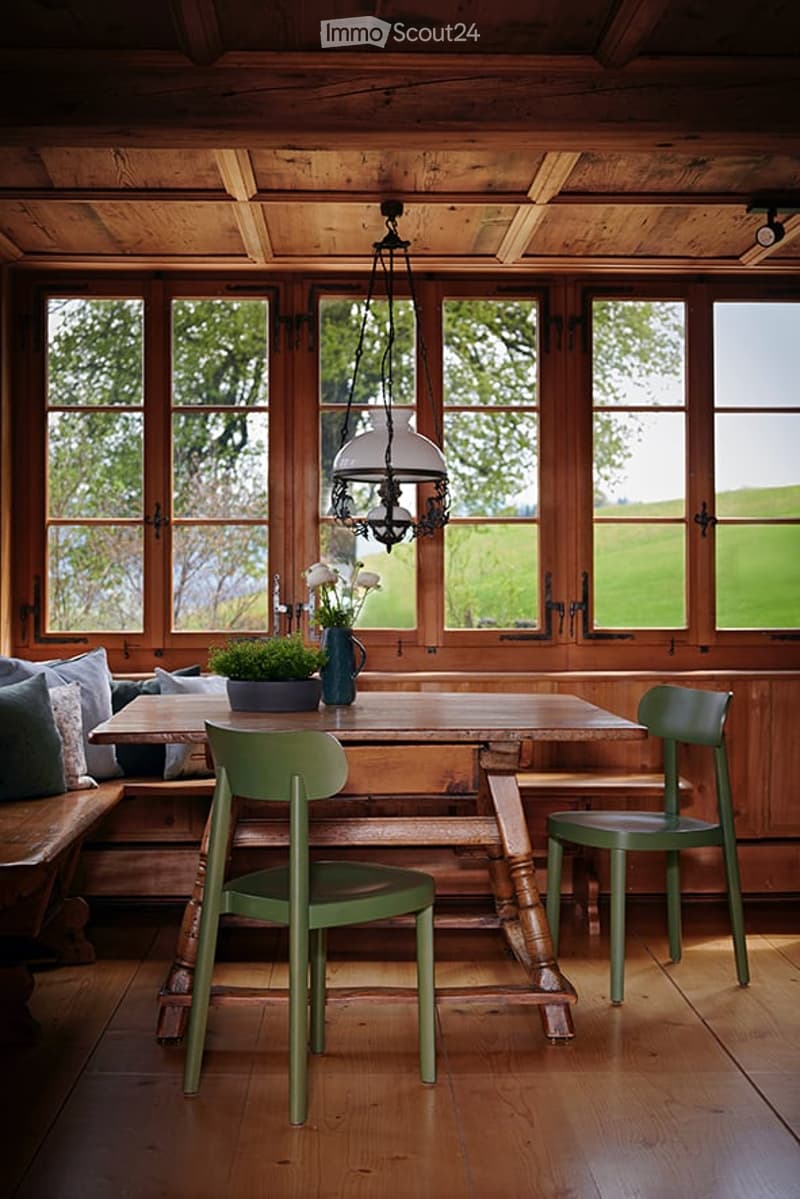 A rustic dining area with wooden table, green chairs, bench seating, and a hanging lantern light fixture. The room features large windows with a view of greenery outside, and a vase with white flowers on the table.