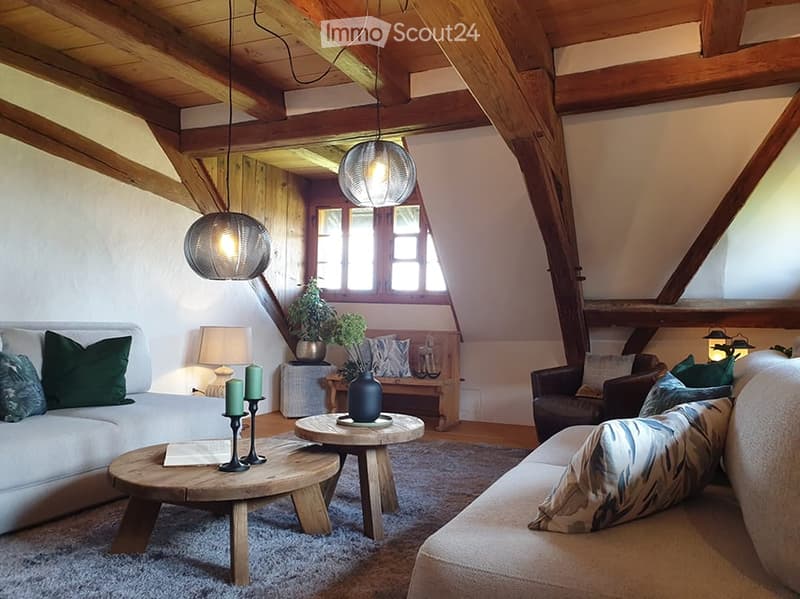Cozy living room with exposed wooden beams, white walls, and large windows. Features a grey sofa, wooden coffee table, floor lamp, and decorative bench with plants.