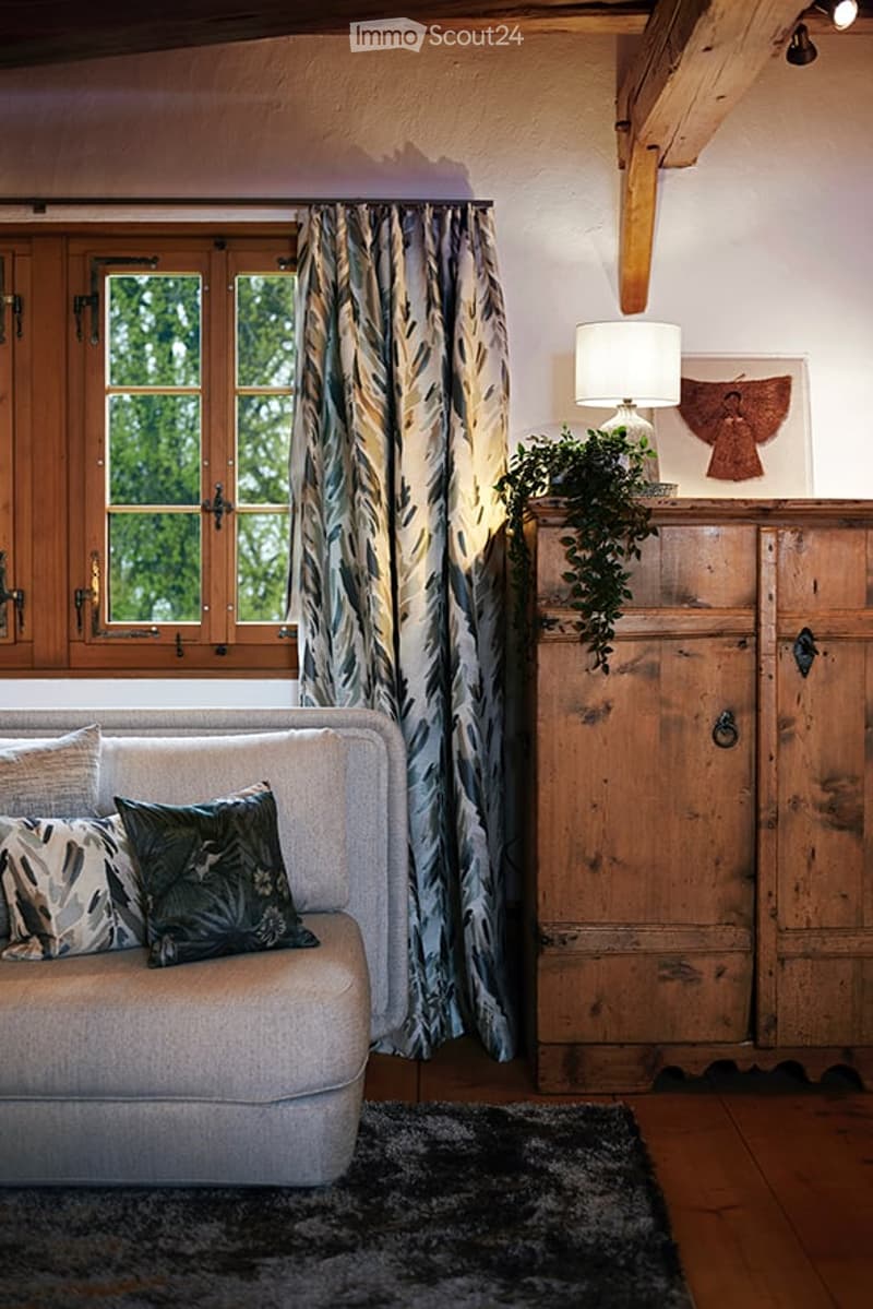 A cozy living room with wooden beams, a large window with patterned curtains, a grey sofa with decorative pillows, a wooden cabinet with a table lamp and plant, and a dark area rug on the floor.