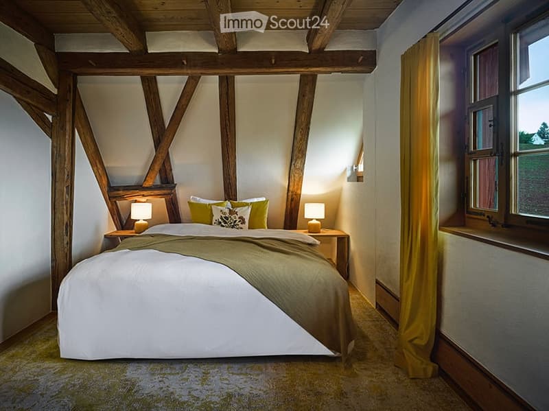 Cozy bedroom with exposed wooden beams, a large bed with green and white bedding, nightstands with lamps, and a window with yellow curtains.