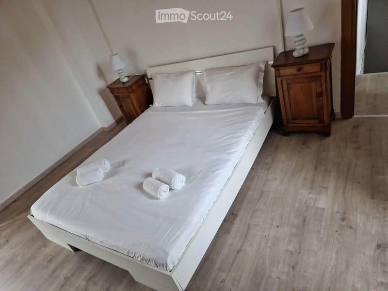 A neatly made double bed with white linens and four rolled towels placed on top, flanked by two wooden nightstands with lamps.