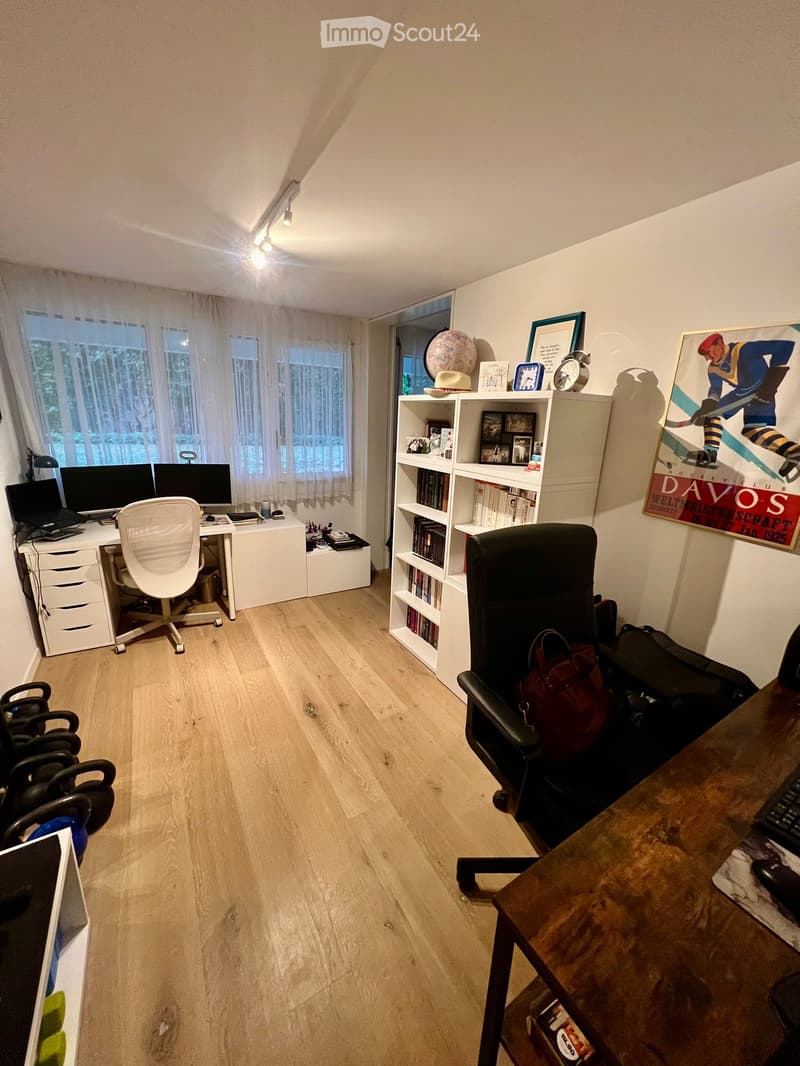 A well-lit room with wooden flooring, a desk with a laptop and screen, a white chair, a bookshelf with books and decorative items, and a large window with curtains.