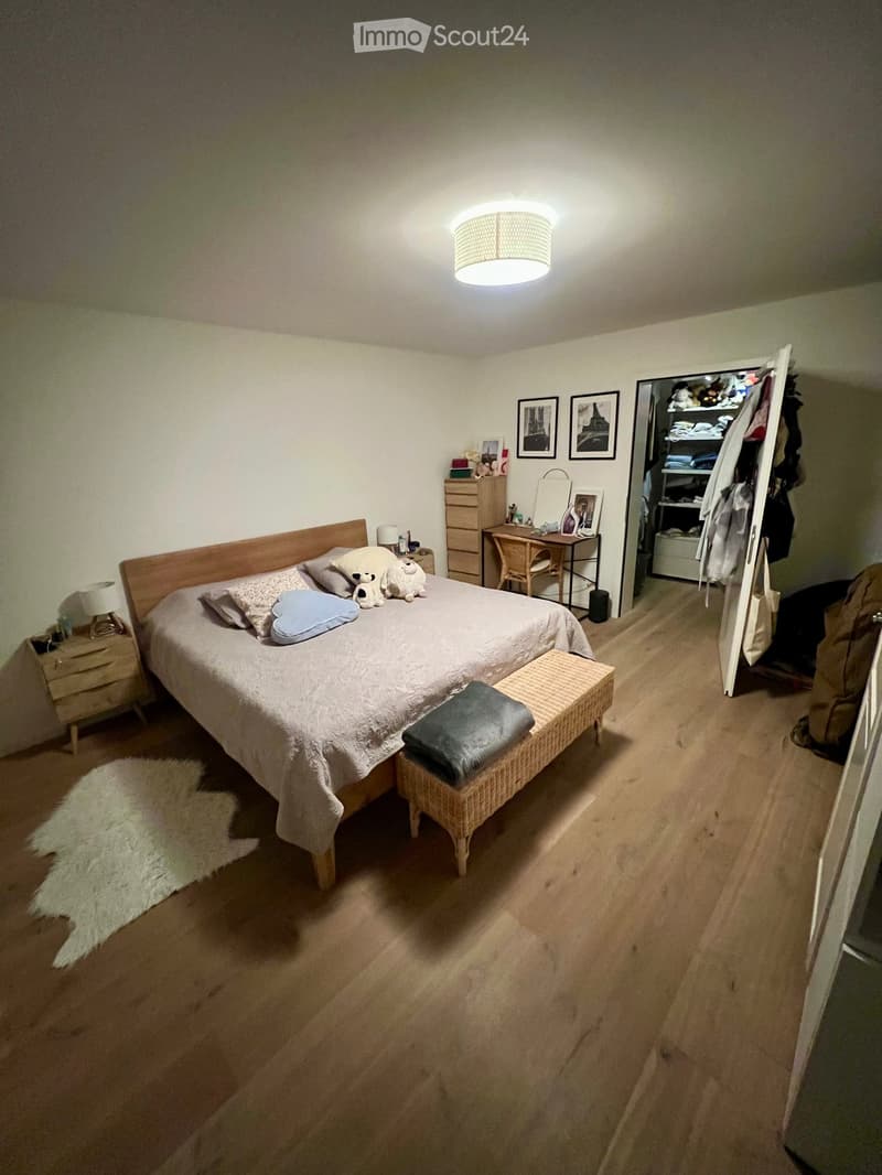 Cozy bedroom with wooden flooring, a large bed with pillows and a blanket, a nightstand with a lamp, a wicker stool, a small desk with a chair, and an open wardrobe with clothes and storage.