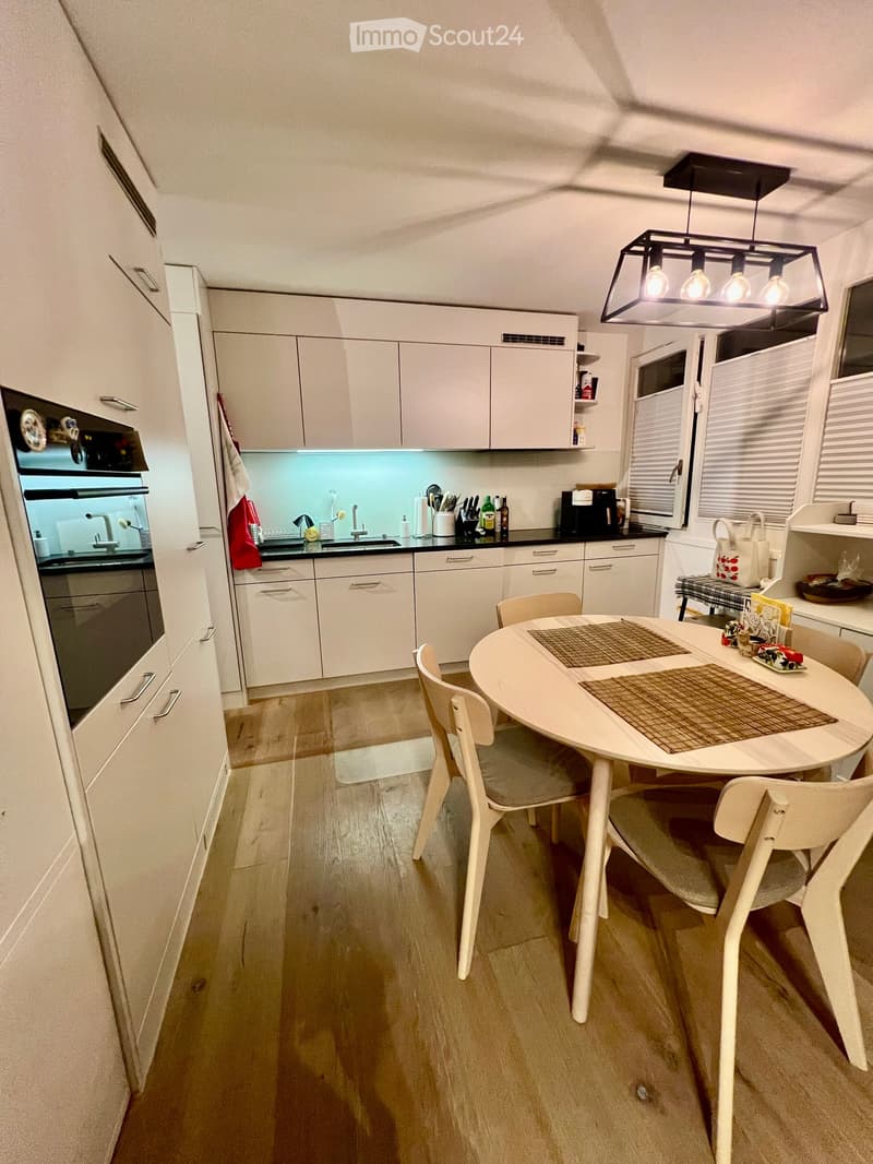 Modern kitchen with white cabinets, a round dining table with four chairs, and various appliances including an oven, coffee maker, and sink.
