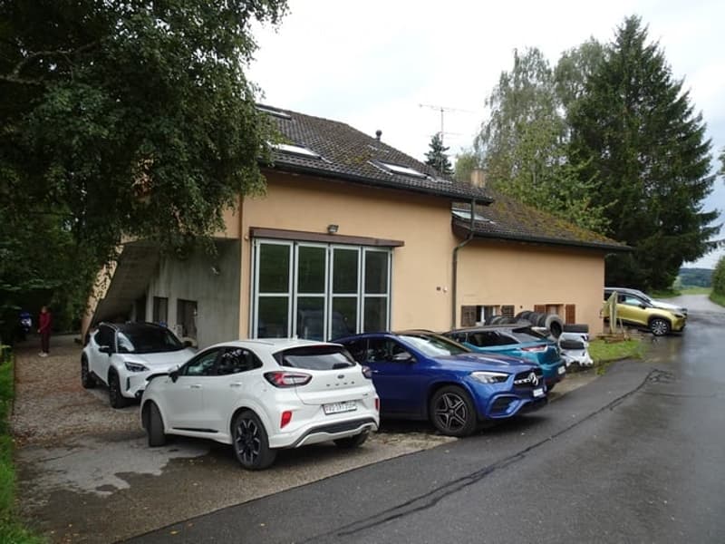 A modern two-story building with a garage and several parked cars in front. The building has large windows and a sloped roof.