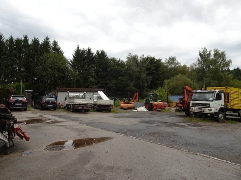 Outdoor area with several parked vehicles, including trucks and cars, surrounded by trees and a small building in the background.