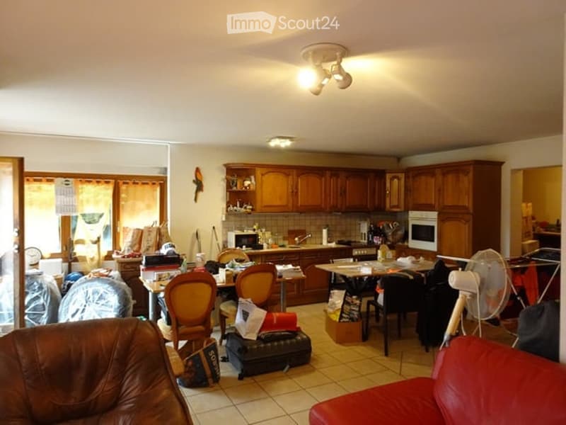 The room is furnished with a leather sofa, wooden chairs, a table, and a fan. There is a kitchen area with wooden cabinets, an oven, and a microwave. The room has tiled flooring and a ceiling light fixture.