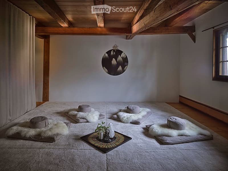Minimalist bedroom with wooden beams, meditation cushions, furs, and a small table with a vase and flower.
