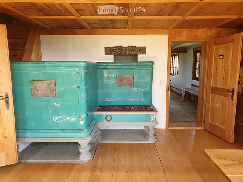 Room with wooden floors and ceiling, featuring a large teal fireplace with decorative metal plates and a small bench visible through an open doorway.