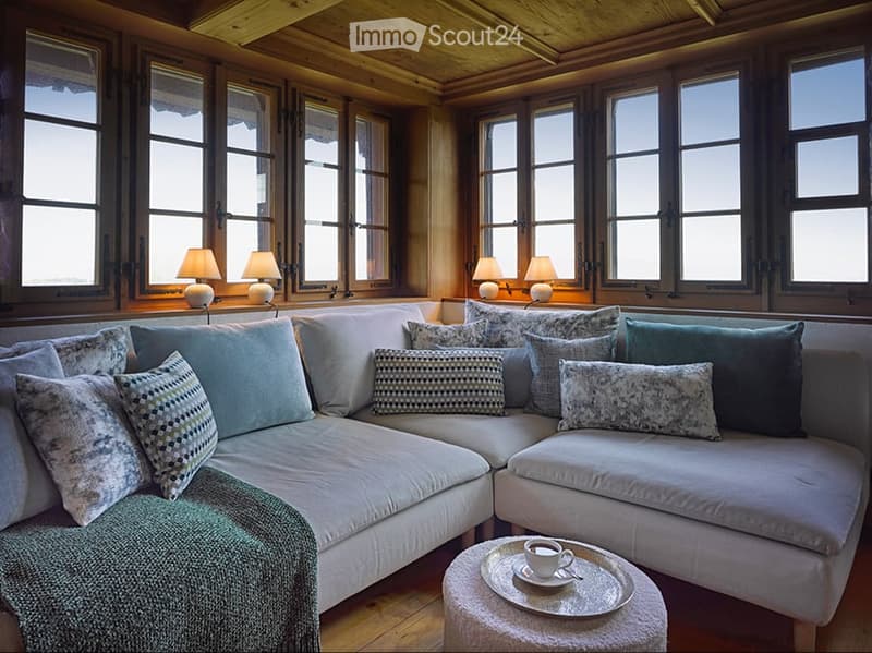 Cozy living room with large windows, wooden ceiling, and a sectional sofa adorned with multiple pillows. A small coffee table holds a cup and saucer.