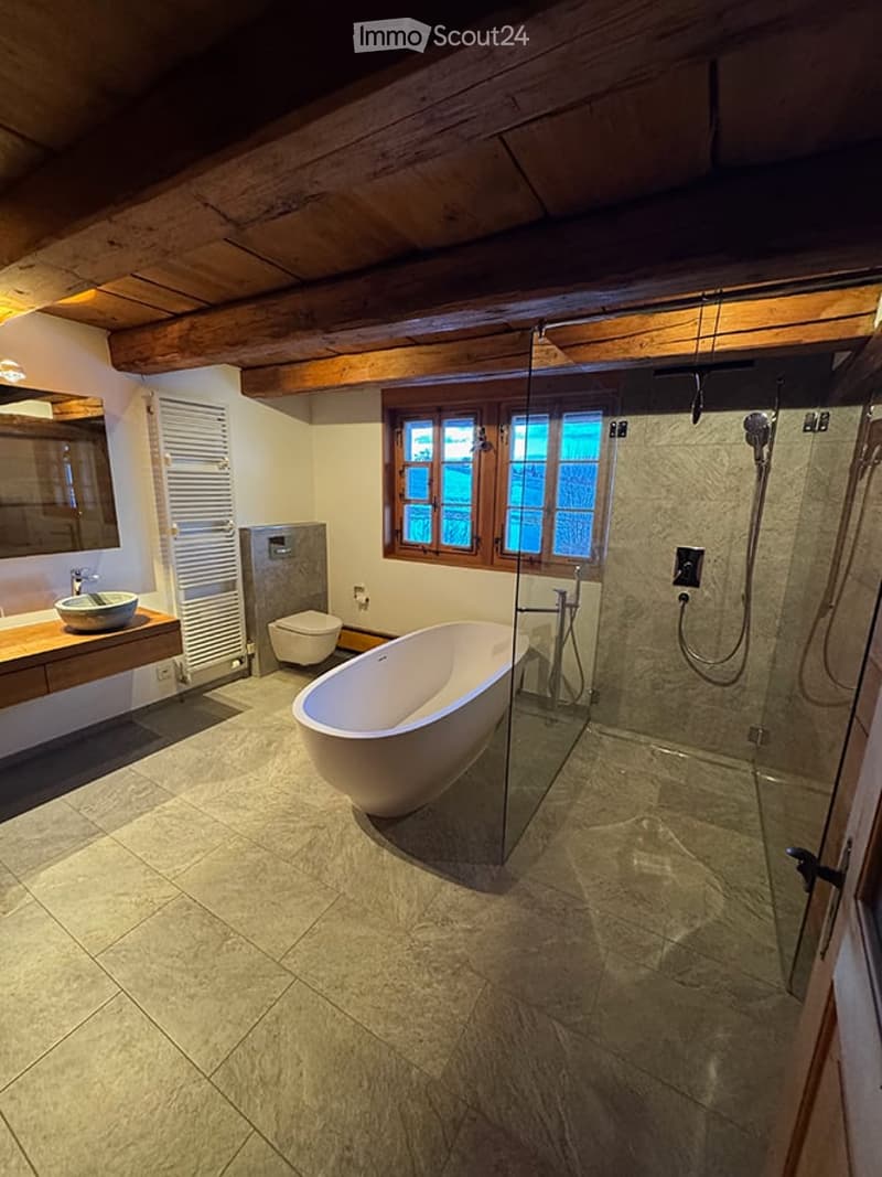 Modern bathroom with a freestanding bathtub, glass-enclosed shower, toilet, sink, and mirror. Exposed wooden beams on the ceiling and large windows provide natural light.