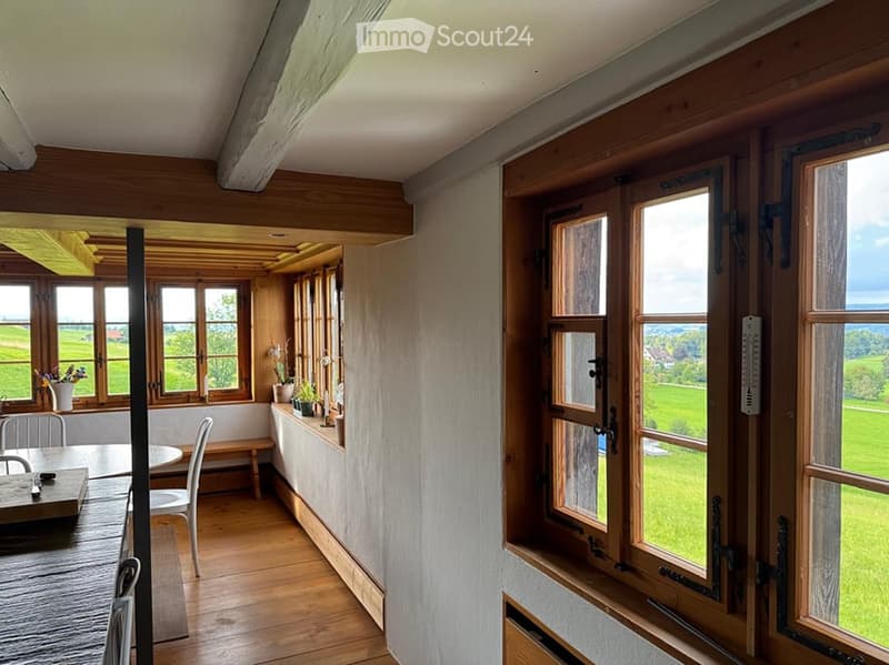 Bright, airy room with wooden floors and large windows overlooking a green field. Features a dining table with chairs, a bench, and several vases with flowers on the windowsill.