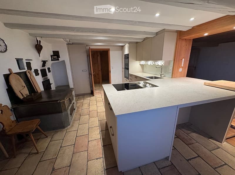 Modern kitchen with a central island, built-in oven, sink, and wooden chair. Exposed wooden beams and tiled floor.