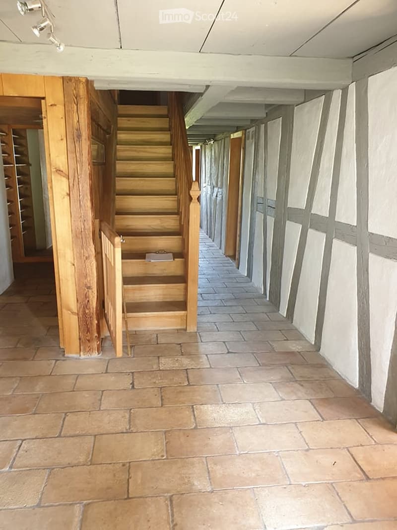 A long, narrow hallway with exposed wooden beams and a wooden staircase leading upwards. The floor is made of terracotta tiles, and the walls are painted in a neutral tone with white trim around the windows. The hallway has several doors on one side, and