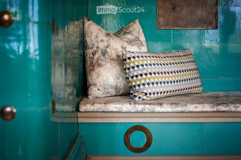 A small bench with two decorative pillows against a teal wall, featuring a metallic drawer below.