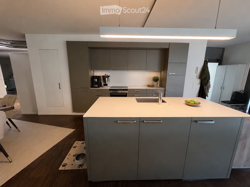 Modern kitchen with grey cabinets, white countertops, and a central island. Features a sink, coffee maker, blender, and a small bowl of fruit on the island.