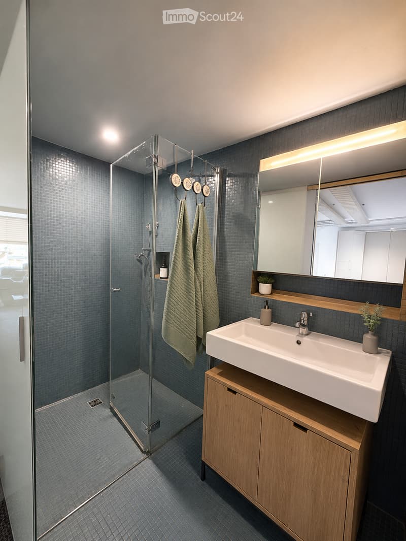 Modern bathroom with a white sink, wooden cabinet, large mirror, glass-enclosed shower, and neatly hung towels.