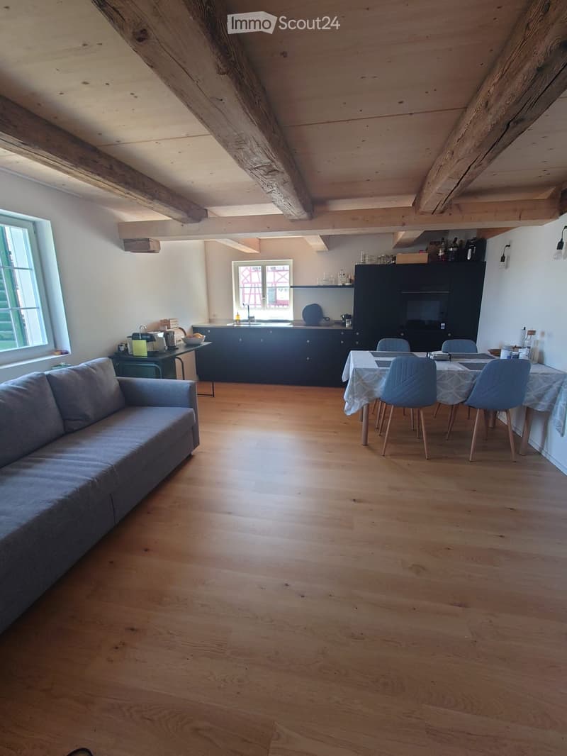 Open-plan living and dining area with exposed wooden beams, hardwood flooring, a grey sofa, a dining table with chairs, and a modern kitchen area with cabinets and appliances.