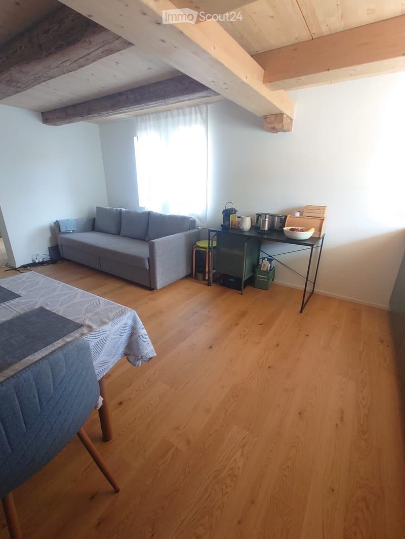 Living room with wooden floor, grey sofa, small black table with various items, and a covered chair.