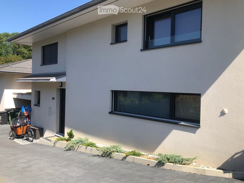 Modern two-story house with light-colored exterior, large windows, and a covered entrance area. Features include a paved driveway, small garden beds, and a parked tricycle.