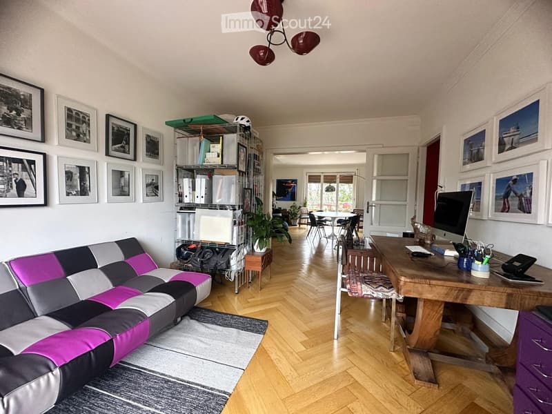 Living room with a modern sofa, wooden floor, large windows, desk with computer, and shelves with various items.