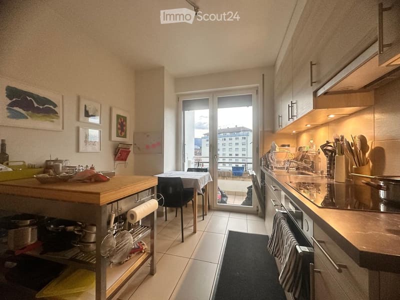 Modern kitchen with a central island, built-in appliances, and a view of the city through a large window. The space includes a dining area with a table and chairs, and various kitchen utensils and decor are visible.