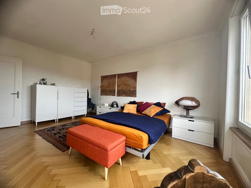 Modern bedroom with wooden flooring, a large bed with colorful pillows and blankets, an orange ottoman, a white wardrobe, and a stylish lamp on a nightstand.