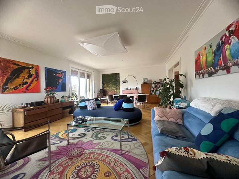 A modern living room with a blue sectional sofa, glass coffee table, colorful rug, and large windows. Features include a dining area with chairs and a table, a floor lamp, and several decorative paintings on the walls.