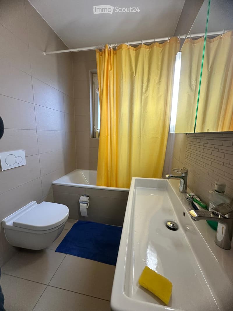 Modern bathroom with a toilet, sink, bathtub, and mirror. Features a yellow shower curtain, a blue bath mat, and a yellow sponge on the sink.