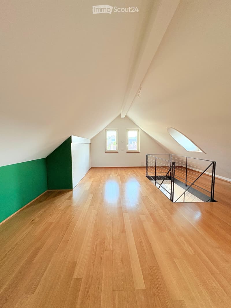 Empty attic room with wooden flooring, white walls, and a metal staircase leading downwards. Features include two windows and a skylight.