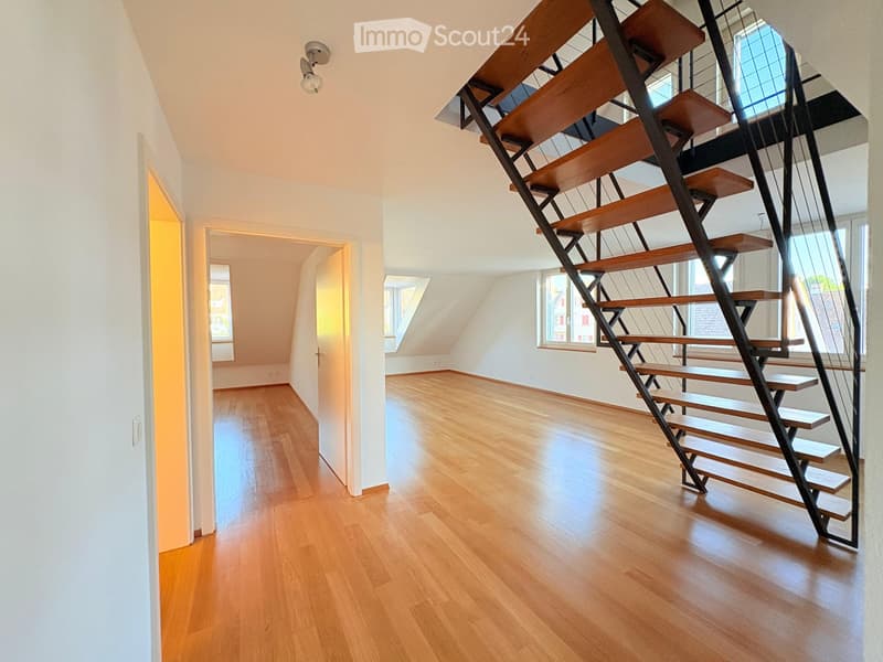 Modern hallway with wooden flooring, white walls, and a contemporary staircase featuring wooden steps and metal railings. Natural light streams in from windows on the right side.