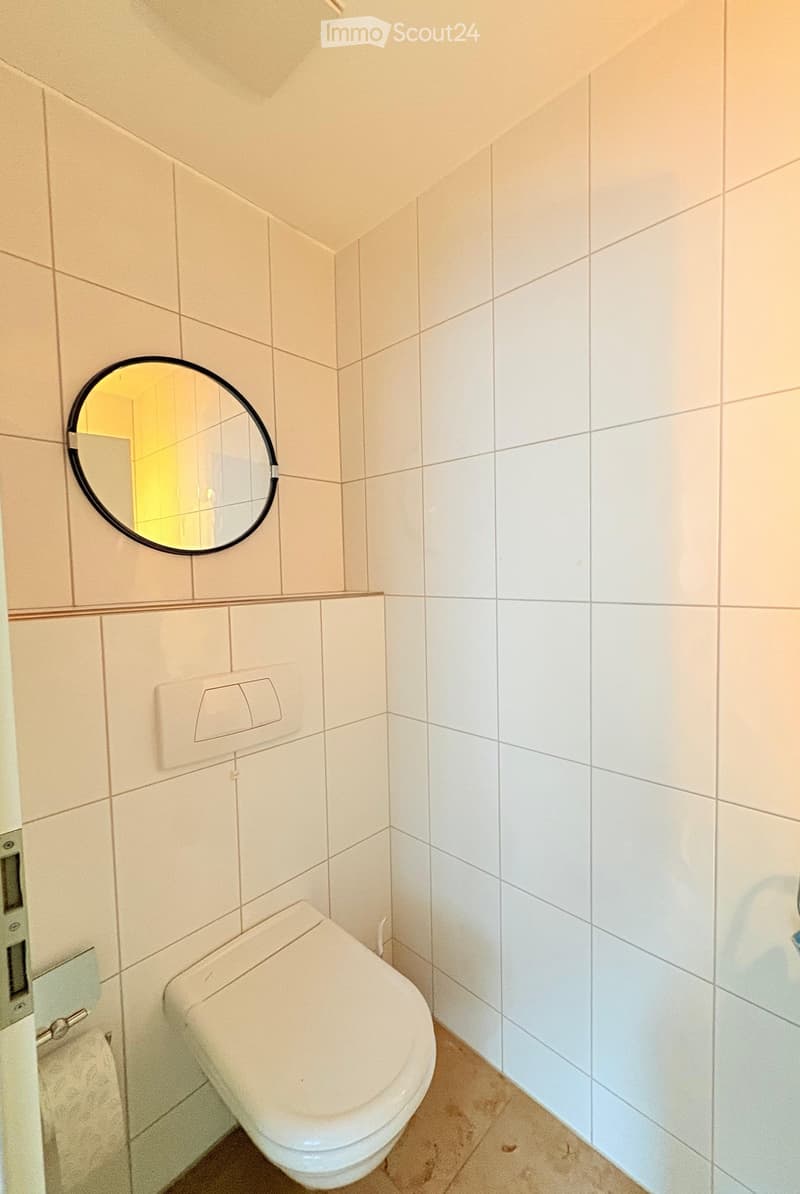 Small bathroom with white tiled walls, a toilet, and a round mirror mounted on the wall.