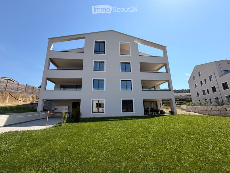 Modern two-story house with large windows and balconies, surrounded by a well-maintained lawn and stone wall.