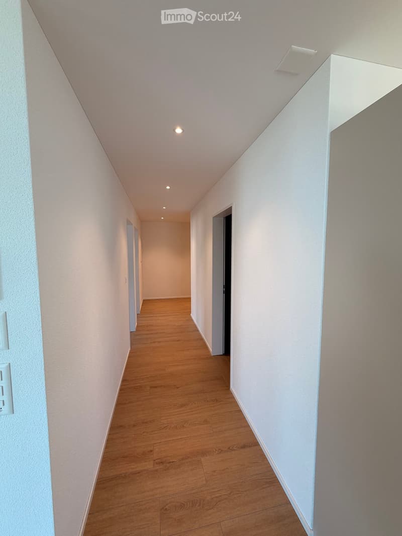 Empty hallway with wooden flooring, white walls, and recessed ceiling lights. Doors are visible at both ends.
