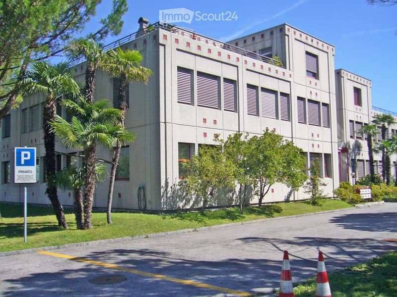 Modern multi-story building with a clean, geometric facade, surrounded by palm trees and greenery. A parking sign is visible in the foreground.