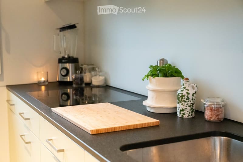 Modern kitchen with white cabinets, black countertop, and various items including a blender, cutting board, potted plant, bottle, jar, and candle.
