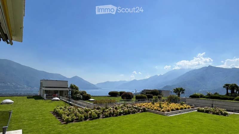 Well-maintained garden with neatly trimmed hedges, flower beds, and a small building, overlooking a scenic lake and mountain range.