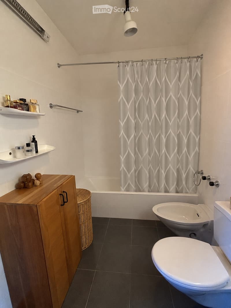 Modern bathroom with dark tiled floor, white walls, wooden cabinet, wicker basket, shelves with toiletries, bathtub, toilet, and shower curtain.