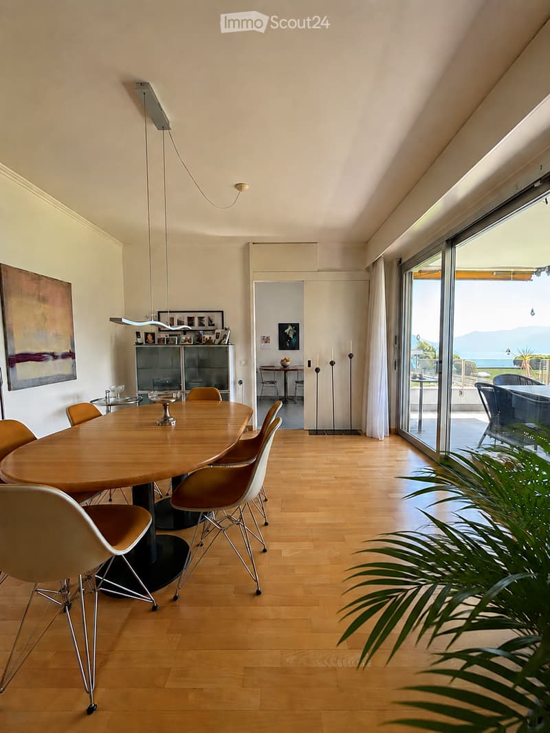 Bright, modern dining area with a round wooden table, chairs, a cabinet with glass doors, and a view of the sea through large windows.