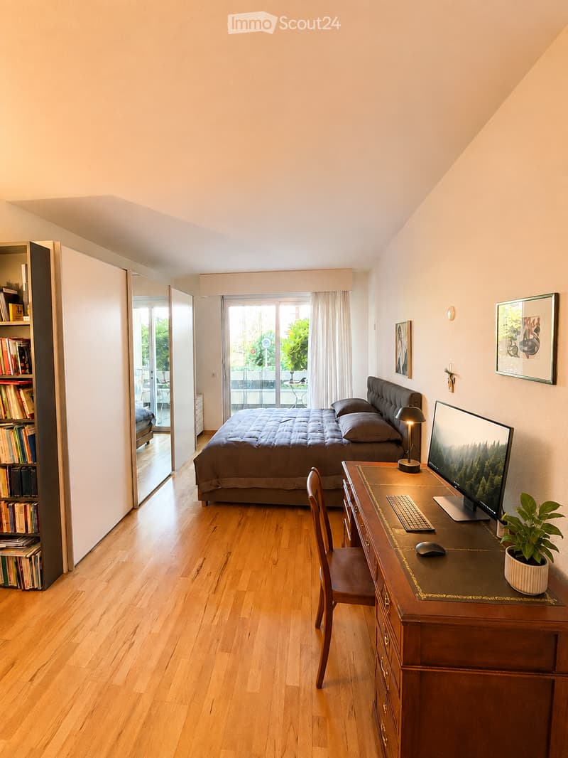 Bright bedroom with wooden flooring, large bed with dark bedding, desk with computer setup, bookshelf, and large window with view to balcony.