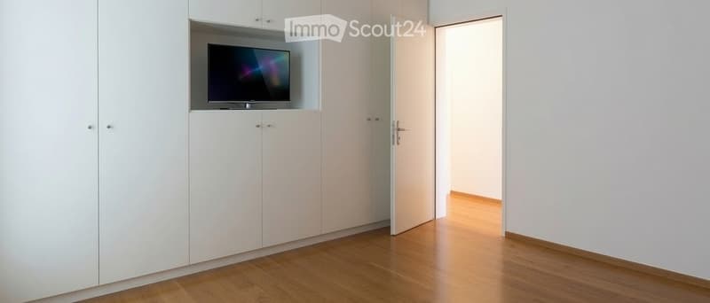 A modern, minimalist room with light wooden flooring, white walls, and built-in white cabinetry. A flat-screen TV is mounted on the cabinetry, and an open doorway leads to another space.