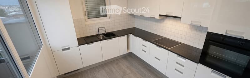 Modern kitchen with white cabinets, black countertops, a sink, stove, and oven. Large window offers a view of the cityscape.