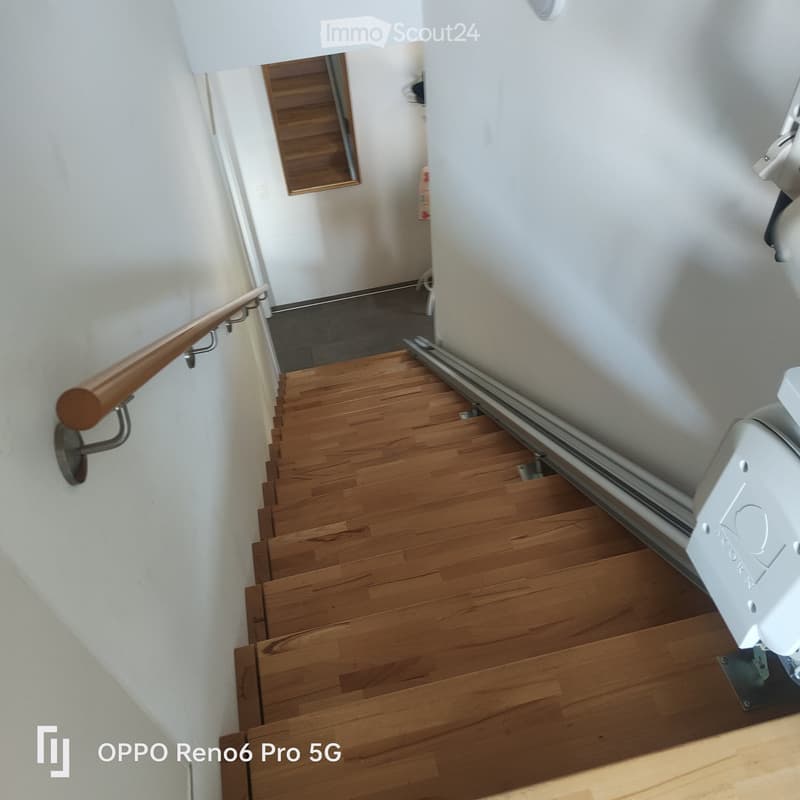 Wooden staircase with a polished finish, white walls, and a metal handrail on one side. The stairs lead upwards, and a small section of a hallway is visible at the top.