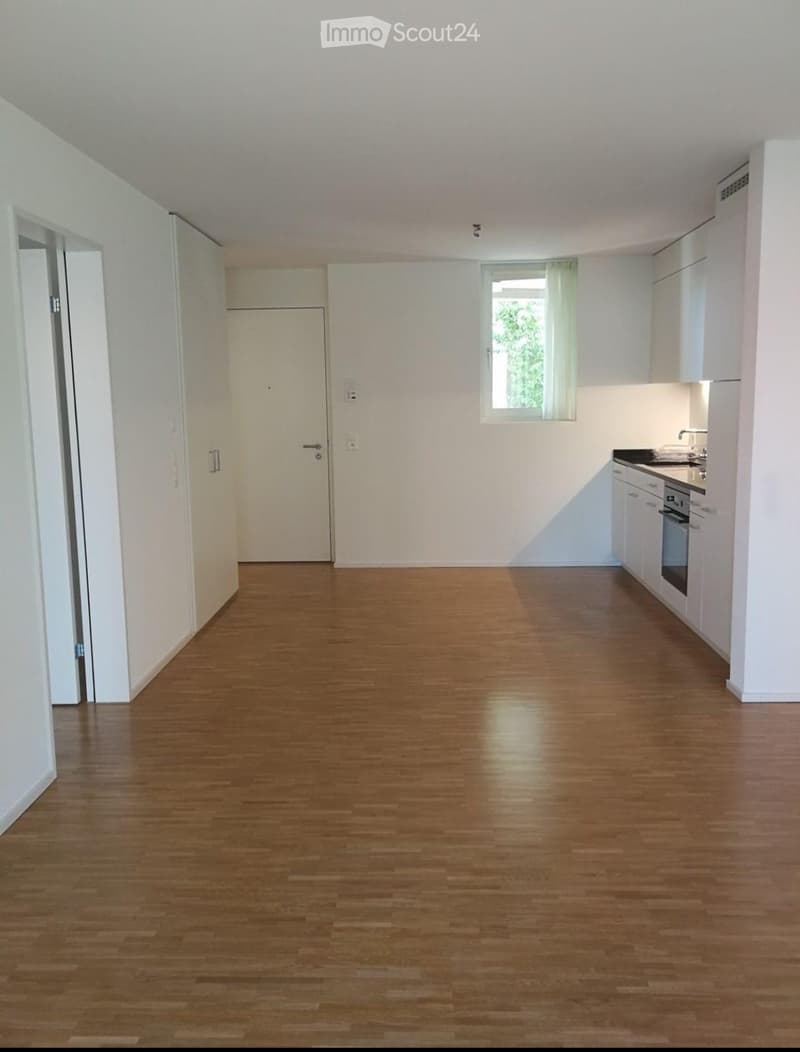 Empty room with wooden flooring, white walls, and a small kitchen area with cabinets and appliances visible in the background.