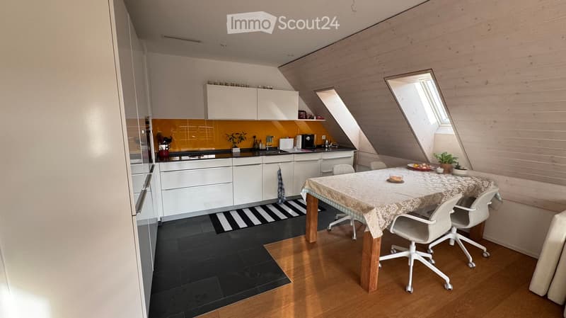A modern kitchen and dining area with white cabinets, a black countertop, a dining table with chairs, and a small plant on the table. The room features a slanted wooden ceiling and large windows.