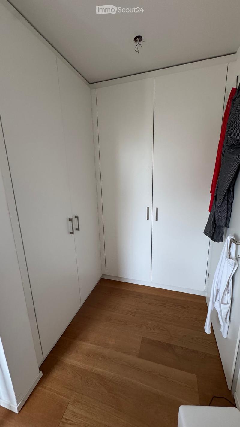 Empty room with white walls, wooden floor, built-in white wardrobes, and a coat rack with a few hanging items.