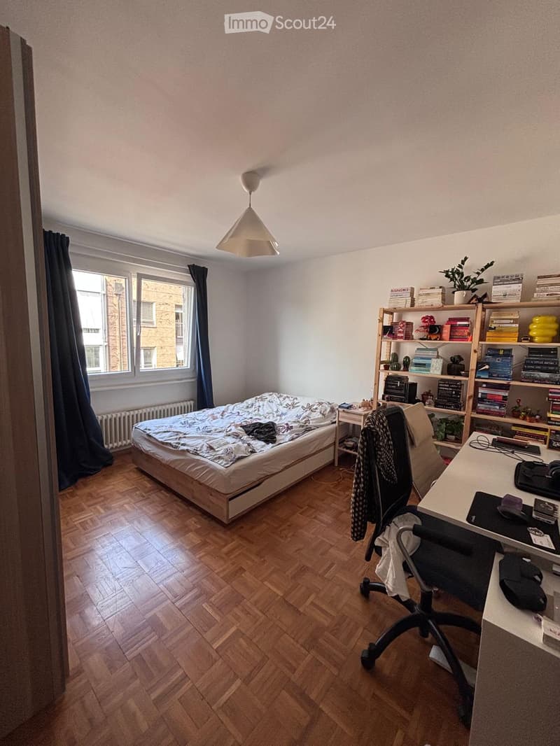 Cozy bedroom with wooden flooring, a bed with a patterned duvet, a desk with a chair, a bookshelf filled with books and decorative items, and a hanging lamp.
