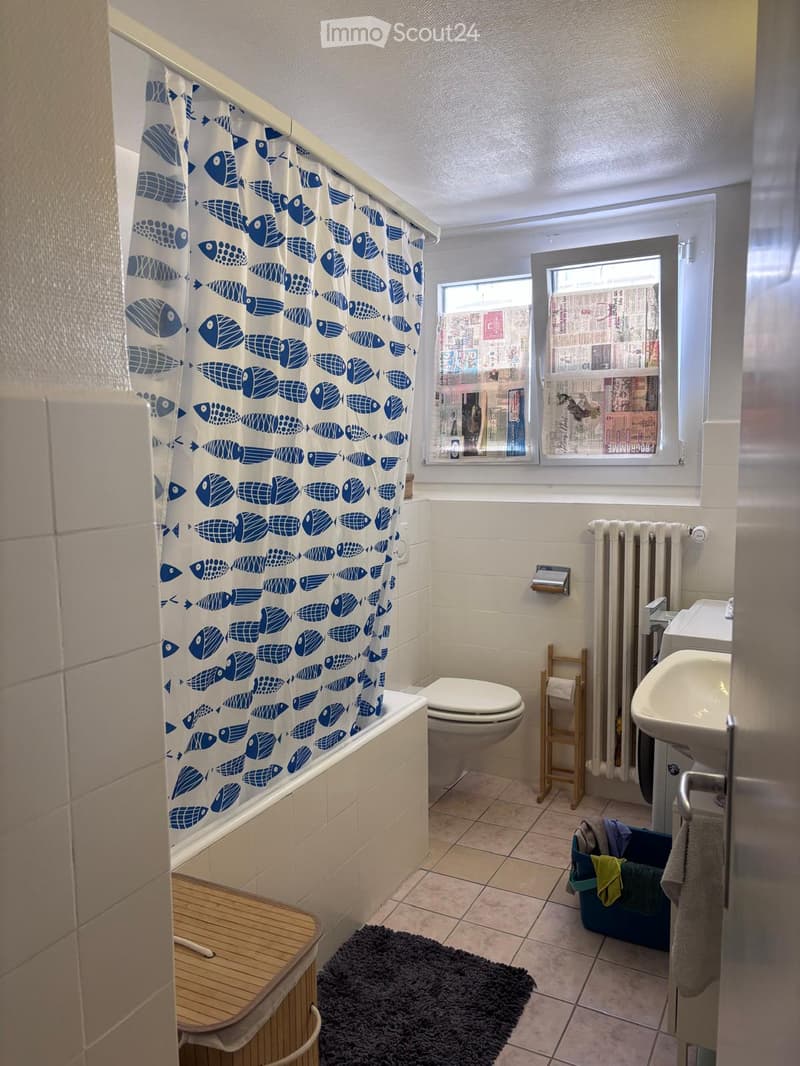 Bathroom with a bathtub, toilet, sink, radiator, and a small wooden stool. A blue fish-patterned shower curtain is visible, along with a towel and a rug on the floor.