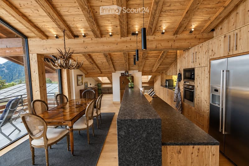A modern, open-concept kitchen and dining area with exposed wooden beams, a large island, and a dining table with chairs. The space features stainless steel appliances, wooden cabinets, and a chandelier made of antlers.