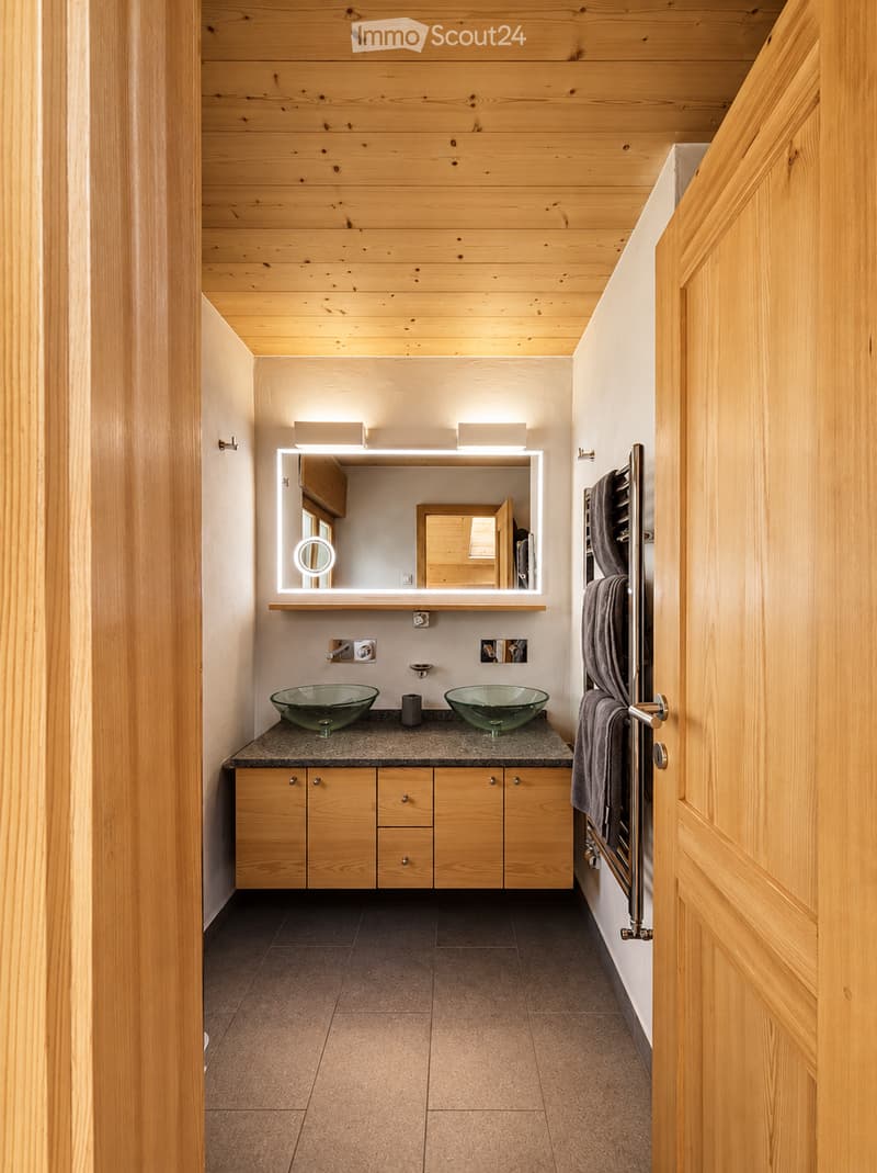 Modern bathroom with wooden walls and ceiling, two glass sinks, a large illuminated mirror, and a towel rack with a towel.
